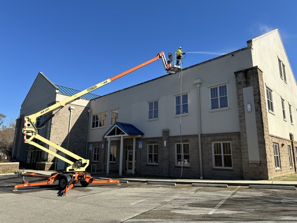 The image contains a large commercial building that is being cleaned by the Water Works crew. There is a crew member cleaning the roof near the roofline thanks to a large crane helping him up there. He's spraying water on the building.